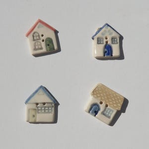 May include: Four ceramic buttons shaped like small houses. Each button features two holes for sewing. The designs include a house with a red roof and green door, a blue roof with a blue door, a blue roof with a light door, and a tan roof.