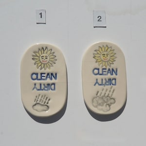 May include: Two ceramic dishwasher magnets. Each has a smiling yellow sun at the top, with the words "CLEAN" and "DIRTY" in blue. One shows rain, the other shows a cloud. The magnets are off-white and oval-shaped.