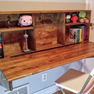 Könnte beinhalten: Ein ausklappbarer Holzschreibtisch mit Regalen. Der Schreibtisch ist an der Wand montiert und hat eine helle Holzoptik. Die Regale sind mit Büchern, Spielzeug und einem kleinen Modell des Space Needle gefüllt.