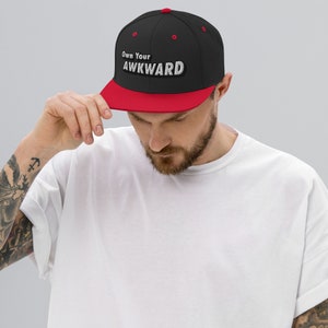 Può includere: Un cappello da baseball nero e rosso con il testo "Own Your AWKWARD" in lettere bianche.