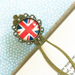 Puede incluir: Un marcapáginas de estilo vintage con un diseño de la bandera británica. El emblema redondo, con colores rojo, blanco y azul, está engastado en un marco de filigrana de tono bronce con detalles florales. El marcapáginas está en un libro.