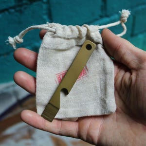 May include: A brass bottle opener and pry bar tool with a small hole at the top and a notch for opening bottles. The tool is displayed with a small, off-white drawstring bag with a red stamp.