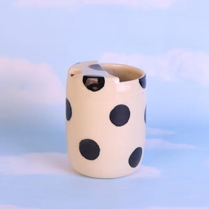 May include: A cream ceramic vase decorated with large black polka dots. The vase has an unusual, asymmetrical top and a cylindrical form. The background is a light blue sky with fluffy white clouds.