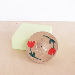 May include: A beige ceramic incense burner with a small hole in the centre. The burner is decorated with two red tulips and green leaves.