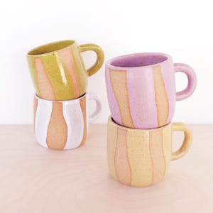 May include: A white ceramic mug with a black handle and the text "But First, Coffee" in a bold, black font. The mug is set against a blurred background. The mug appears to be approximately 10 cm tall.