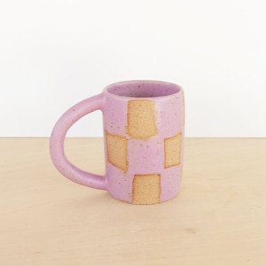May include: A purple ceramic mug with a geometric pattern of tan squares. The mug has a rounded handle.