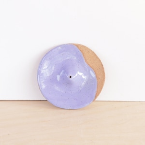 Peut inclure: Un porte-encens rond en céramique avec un glaçage lavande moucheté et une section en argile naturelle. Il comporte un petit trou central pour les bâtonnets d'encens. Design minimaliste et moderne, environ 10 cm de diamètre.
