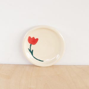 May include: A small, round, cream-coloured ceramic dish featuring a hand-painted red tulip design. The tulip has a green stem and is centrally placed. This dish is suitable for small items or as a decorative element.
