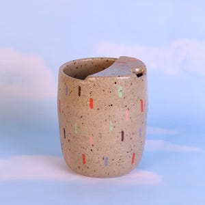 Peut inclure: Une tasse en céramique tachetée avec un repose-couvercle intégré. La tasse est beige avec des tirets verticaux colorés en rouge, rose, vert et bleu. Le fond est un ciel bleu clair avec des nuages blancs.