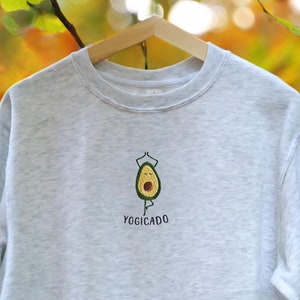 Op de afbeelding: Een lichtgrijs sweatshirt met een groene avocado die yoga doet en de tekst "Yogicado" geborduurd op de voorkant.
