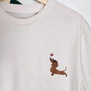 Op de afbeelding: Crèmekleurig T-shirt met een geborduurd bruin teckel dat een wijnglas vasthoudt. De hond is afgebeeld in een speelse pose, met het glas omhoog. Het shirt hangt aan een houten hanger.
