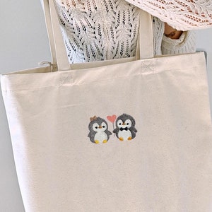 Puede incluir: Un bolso de tela de color natural con un dise&ntilde;o bordado de dos peque&ntilde;os ping&uuml;inos. Un ping&uuml;ino lleva un peque&ntilde;o lazo en la cabeza y el otro una pajarita negra. Entre ambos, sostienen un globo rosa en forma de coraz&oacute;n sujeto por una cuerda.