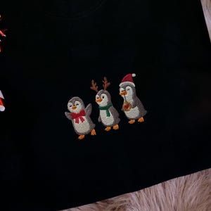 Op de afbeelding: Een zwart sweatshirt met een geborduurd ontwerp van drie pinguïns met feestelijke accessoires. Een zuurstokornament hangt links. Kerstboomtakken en -ballen zijn op de achtergrond te zien.