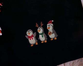 May include: A black sweatshirt featuring an embroidered design of three penguins wearing festive holiday accessories. A candy cane ornament hangs to the left of the sweatshirt. Christmas tree branches and ornaments are in the background.