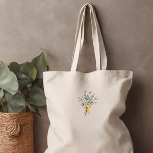 May include: A cream-colored canvas tote bag with a floral embroidery design. The embroidery features a bouquet of white flowers with green leaves and a yellow ribbon.
