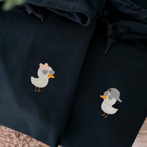 Op de afbeelding: Twee donkerblauwe hoodies met geborduurde eendenontwerpen. Eén eend draagt een zonnebril en een strik, de andere een pet. De hoodies hebben trekkoorden en worden gepresenteerd op een zacht, lichtbruin oppervlak.