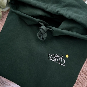 Puede incluir: Una sudadera con capucha de color verde bosque oscuro que presenta un pequeño contorno bordado en blanco de una bicicleta en el pecho. El diseño incluye un diminuto sol amarillo bordado sobre la bicicleta y cordones verdes a juego.