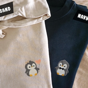 Könnte beinhalten: Zwei Sweatshirts, eines in Sandfarbe und eines in Marineblau, jeweils mit einem Pinguin bestickt. Das sandfarbene Sweatshirt hat einen Pinguin, der einen herzförmigen Ballon hält. Das marineblaue Sweatshirt hat einen Pinguin mit Fliege. Die Wörter "SAND" und "NAVY" sind aufgedruckt.
