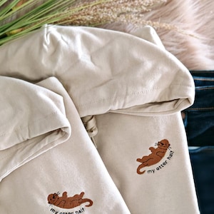 Op de afbeelding: Twee crèmekleurige hoodies met een bruine otter-afbeelding en de tekst "my otter half" geborduurd op de voorkant.
