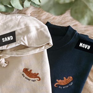 Può includere: Due felpe con un designo di lontra ricamato. Una è una felpa con cappuccio color sabbia con il testo "my otter half" e l'altra è una felpa a collo rotondo blu navy con lo stesso design e testo.