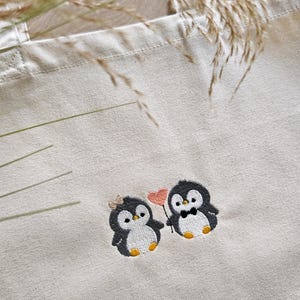 Puede incluir: Una bolsa de tela beige con un dise&ntilde;o bordado de dos peque&ntilde;os ping&uuml;inos. Un ping&uuml;ino lleva un peque&ntilde;o lazo, mientras que el otro lleva una pajarita. Est&aacute;n colocados uno al lado del otro, con uno sosteniendo un palo fino con un coraz&oacute;n rosa.