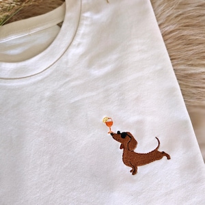 May include: White t-shirt featuring an embroidered brown dachshund wearing sunglasses, balancing a cocktail. The dog is depicted in a playful pose, with the drink held up towards its nose. The shirt is made of a soft, light-colored fabric.