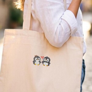 Puede incluir: Una bolsa de tela color beige con un gr&aacute;fico bordado de dos peque&ntilde;os ping&uuml;inos de pie uno al lado del otro. Un ping&uuml;ino lleva un peque&ntilde;o lazo marr&oacute;n, el otro una pajarita negra, y est&aacute;n situados debajo de un peque&ntilde;o icono de coraz&oacute;n rosa.