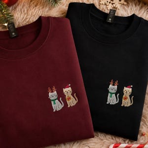 Op de afbeelding: Een bordeauxrood sweatshirt en een zwart sweatshirt, beide voorzien van een borduursel van twee katten. De ene kat draagt een rendiergewei en een groene sjaal, terwijl de andere een rode kerstmuts draagt en een rood hartje vasthoudt.