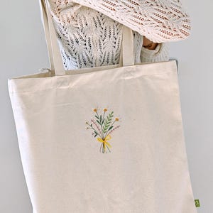 May include: A white canvas tote bag with a small embroidered bouquet of yellow and white flowers tied with a yellow ribbon.