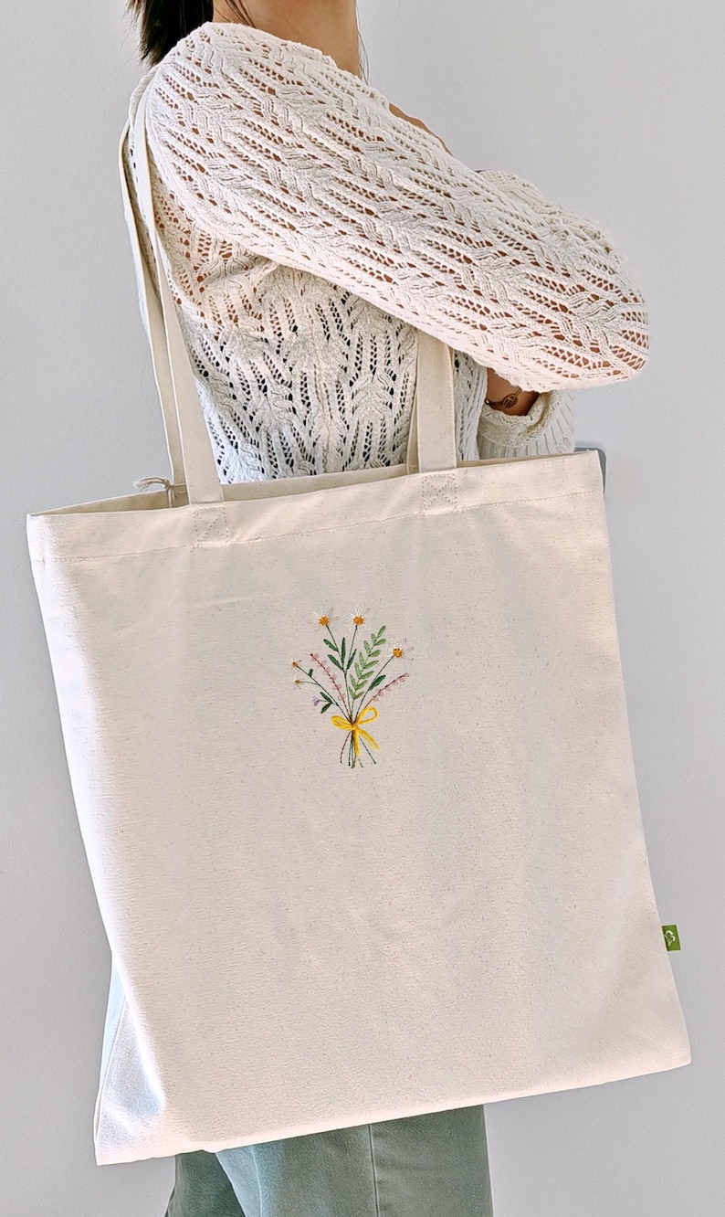 May include: A white canvas tote bag with a small embroidered bouquet of yellow and white flowers tied with a yellow ribbon.
