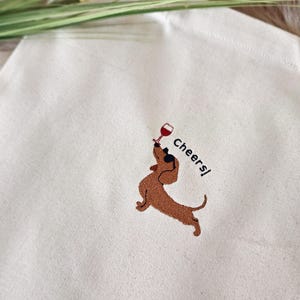 May include: Cream-colored tote bag featuring an embroidered brown dachshund holding a wine glass. The word "Cheers!" is embroidered next to the dog. The bag is made of a light-colored fabric.