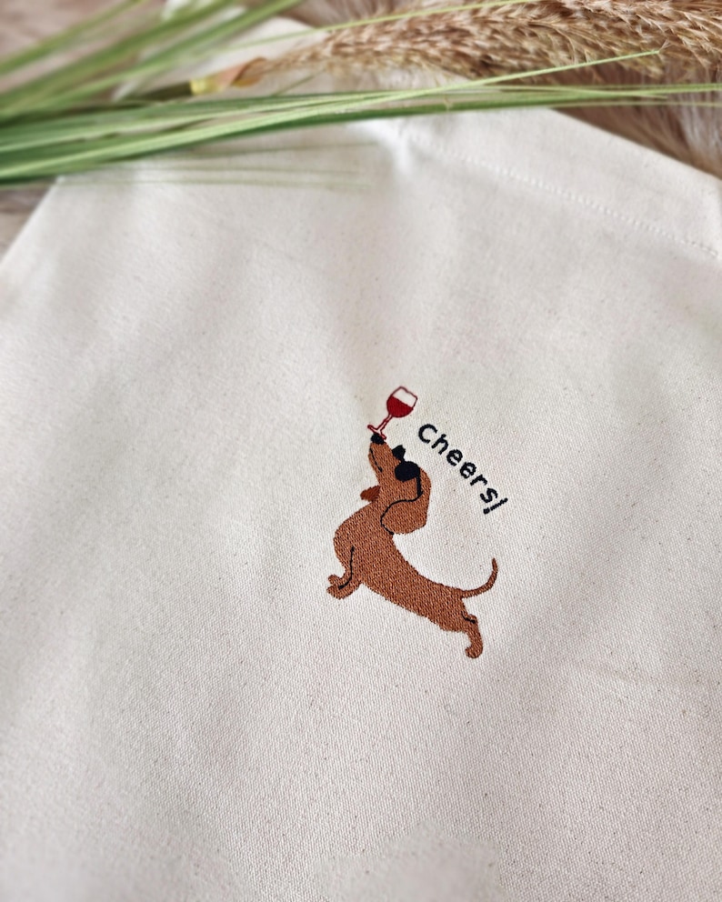 May include: Cream-colored tote bag featuring an embroidered brown dachshund holding a wine glass. The word "Cheers!" is embroidered next to the dog. The bag is made of a light-colored fabric.