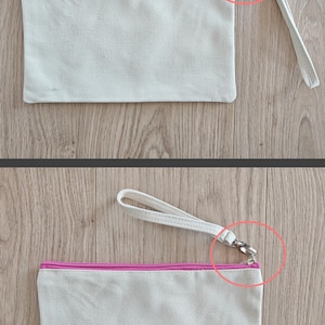 May include: A white canvas zippered pouch with a pink zipper and a white strap with a silver clasp. The pouch is lying flat on a wooden surface.