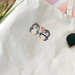 Puede incluir: Un bolso de tela de color blanco roto con un dise&ntilde;o bordado de dos ping&uuml;inos. Un ping&uuml;ino lleva un peque&ntilde;o lazo y el otro una pajarita mientras sostiene un globo rosa en forma de coraz&oacute;n. El bolso tiene dos asas de tela y forma rectangular.