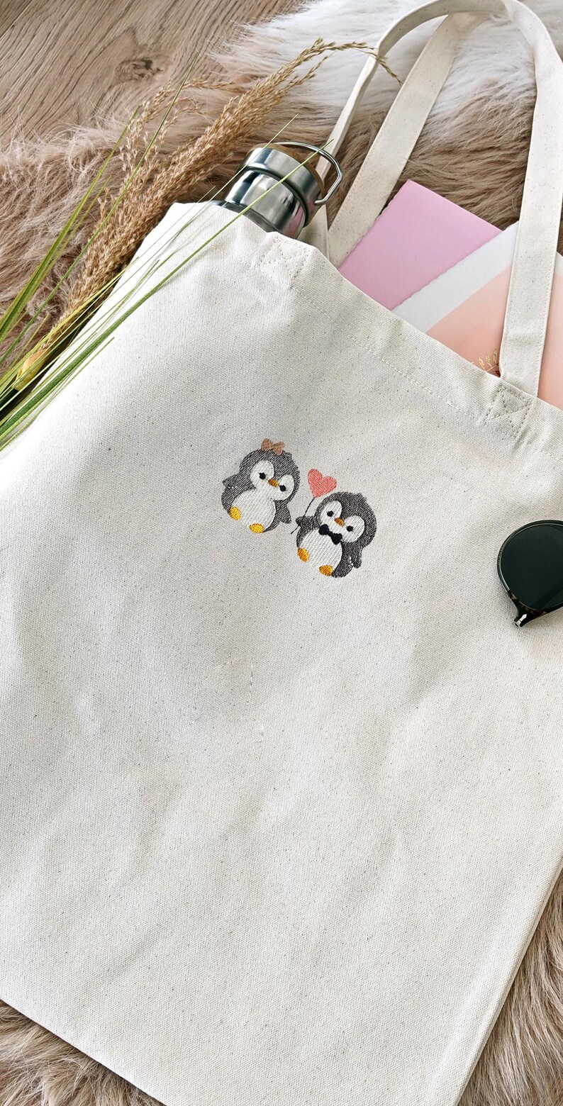 Puede incluir: Un bolso de tela de color blanco roto con un dise&ntilde;o bordado de dos ping&uuml;inos. Un ping&uuml;ino lleva un peque&ntilde;o lazo y el otro una pajarita mientras sostiene un globo rosa en forma de coraz&oacute;n. El bolso tiene dos asas de tela y forma rectangular.
