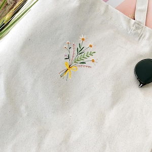 May include: A white canvas tote bag with a small embroidered floral design in yellow, green, and pink. The bag has two long straps.