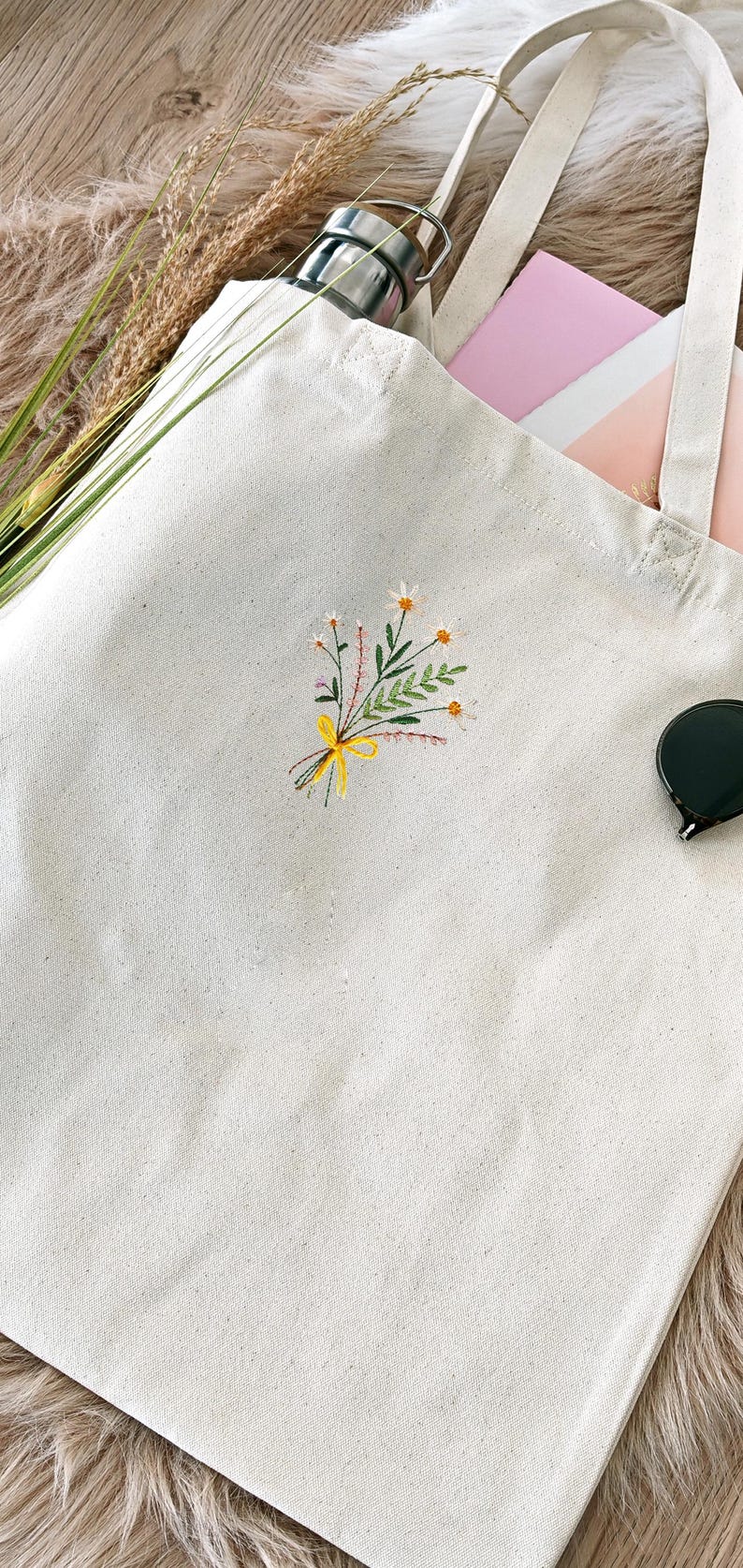 May include: A white canvas tote bag with a small embroidered floral design in yellow, green, and pink. The bag has two long straps.