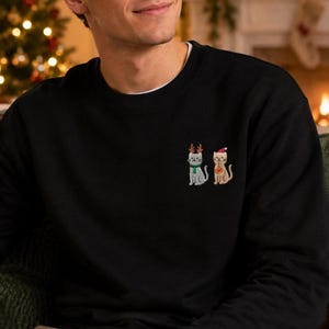 Mann trägt einen Katzen Weihnachts Hoodie mit kleiner Stickerei vor weihnachtlichem Hintergrund.