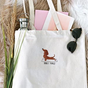 Puede incluir: Una bolsa de lona blanca con un dachshund marr&oacute;n haciendo yoga en una estera y el texto "100% YOGI".