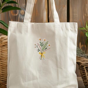 May include: A white canvas tote bag with a small embroidered design of a bouquet of flowers with a yellow ribbon. The flowers are white, pink, and green.