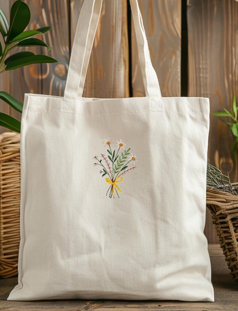 May include: A white canvas tote bag with a small embroidered design of a bouquet of flowers with a yellow ribbon. The flowers are white, pink, and green.