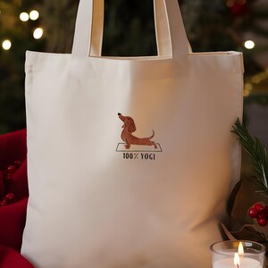 Puede incluir: Bolsa de tela blanca con una imagen bordada de un dachshund haciendo yoga en una colchoneta. El texto "100% YOGI" est&aacute; bordado debajo del dachshund.