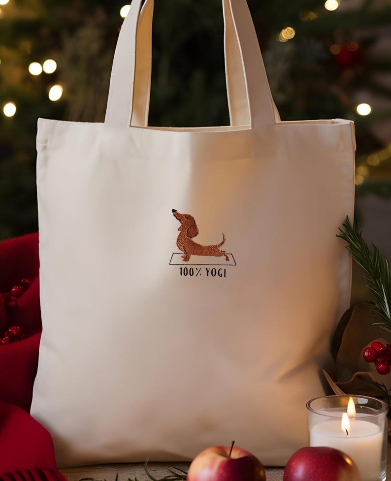 Puede incluir: Bolsa de tela blanca con una imagen bordada de un dachshund haciendo yoga en una colchoneta. El texto "100% YOGI" est&aacute; bordado debajo del dachshund.