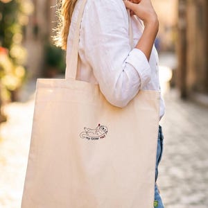 Puede incluir: Un bolso de tela color crema con una peque&ntilde;a ilustraci&oacute;n lineal en negro de una nutria sosteniendo un coraz&oacute;n rojo. Debajo de la nutria, el texto "my Otter Half" aparece escrito en una fuente sencilla y oscura.