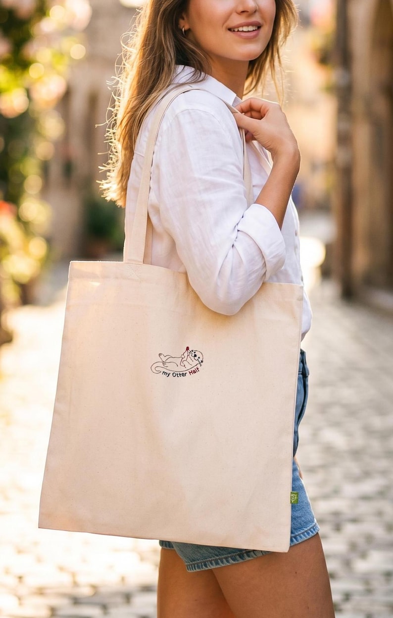 Puede incluir: Un bolso de tela color crema con una peque&ntilde;a ilustraci&oacute;n lineal en negro de una nutria sosteniendo un coraz&oacute;n rojo. Debajo de la nutria, el texto "my Otter Half" aparece escrito en una fuente sencilla y oscura.