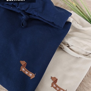 Marineblaue und beige Stoffartikel mit gestickten Dackel-Designs. Der marineblaue Artikel hat einen Kordelzugverschluss. Der Text "EMBROIDERED BESTICKT" ist sichtbar. Die Dackel haben herzförmige Markierungen.
