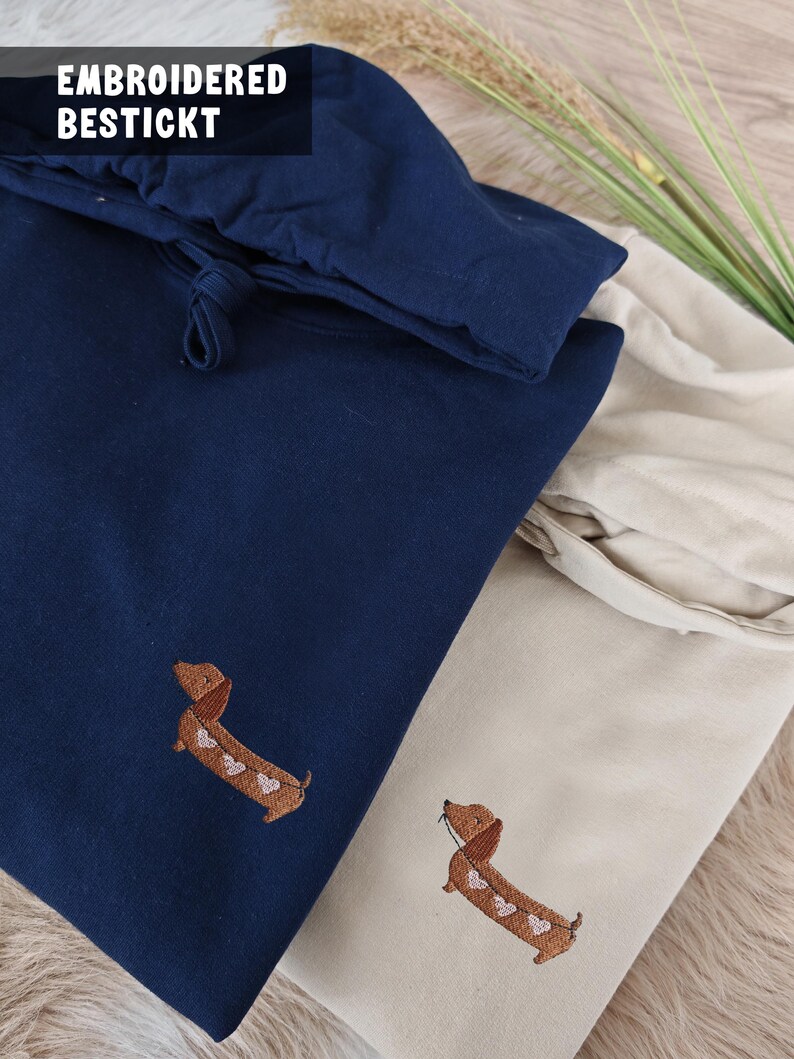 Marineblaue und beige Stoffartikel mit gestickten Dackel-Designs. Der marineblaue Artikel hat einen Kordelzugverschluss. Der Text "EMBROIDERED BESTICKT" ist sichtbar. Die Dackel haben herzförmige Markierungen.