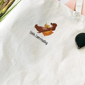 Puede incluir: Una bolsa de lona blanca con un dachshund marr&oacute;n bordado en ella. El dachshund lleva gafas y sostiene un libro. El texto "Shhh. I am reading" est&aacute; bordado debajo del dachshund.