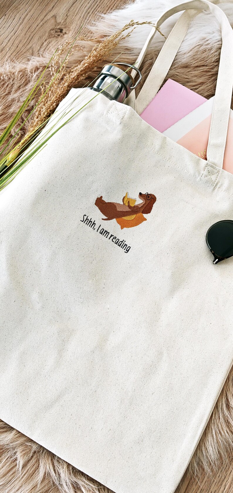 Puede incluir: Una bolsa de lona blanca con un dachshund marr&oacute;n bordado en ella. El dachshund lleva gafas y sostiene un libro. El texto "Shhh. I am reading" est&aacute; bordado debajo del dachshund.