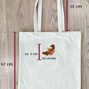 Puede incluir: Una bolsa de tela blanca con un dachshund leyendo un libro. La bolsa tiene asas marrones y mide 38,5 cm de ancho, 42 cm de alto y 30 cm de profundidad. El texto en la bolsa dice "Shhh, I am reading."